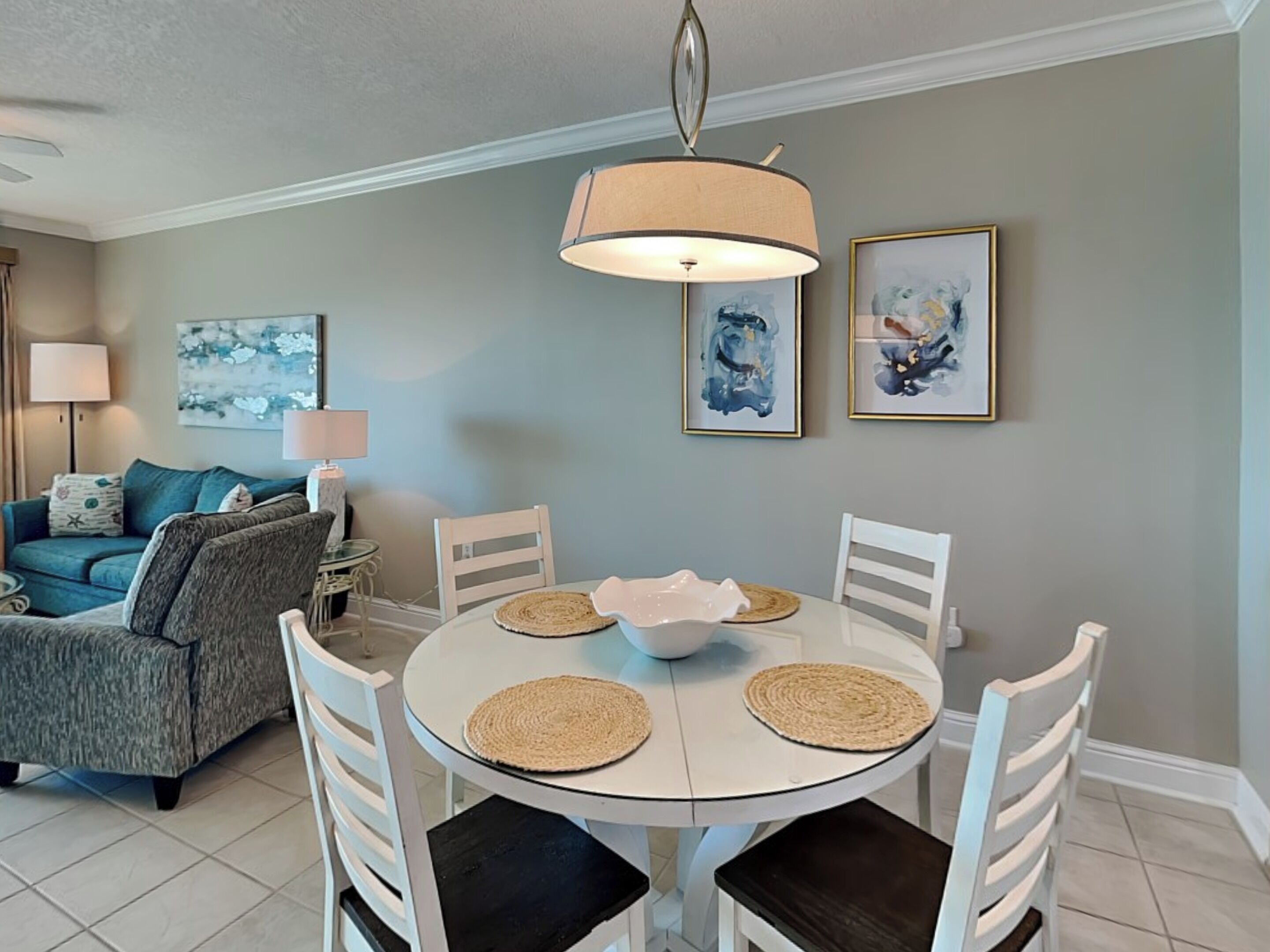 A dining area with a round table, white chairs, and a hanging lamp. There are two framed abstract paintings on the wall.