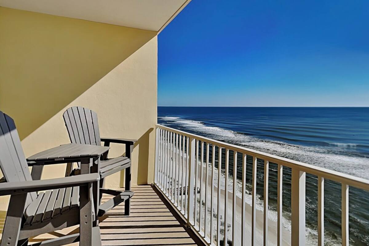 A balcony with a view of the ocean, featuring two Adirondack chairs and a railing.