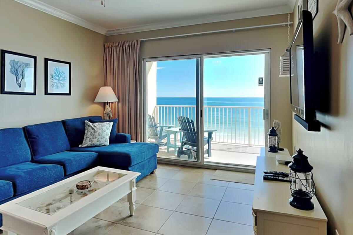 A living room with a blue sofa, a white coffee table, a ceiling fan, and a view of the ocean through a sliding glass door.