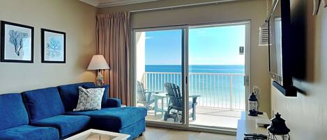 A living room with a blue sofa, a white coffee table, a ceiling fan, and a view of the ocean through a sliding glass door.