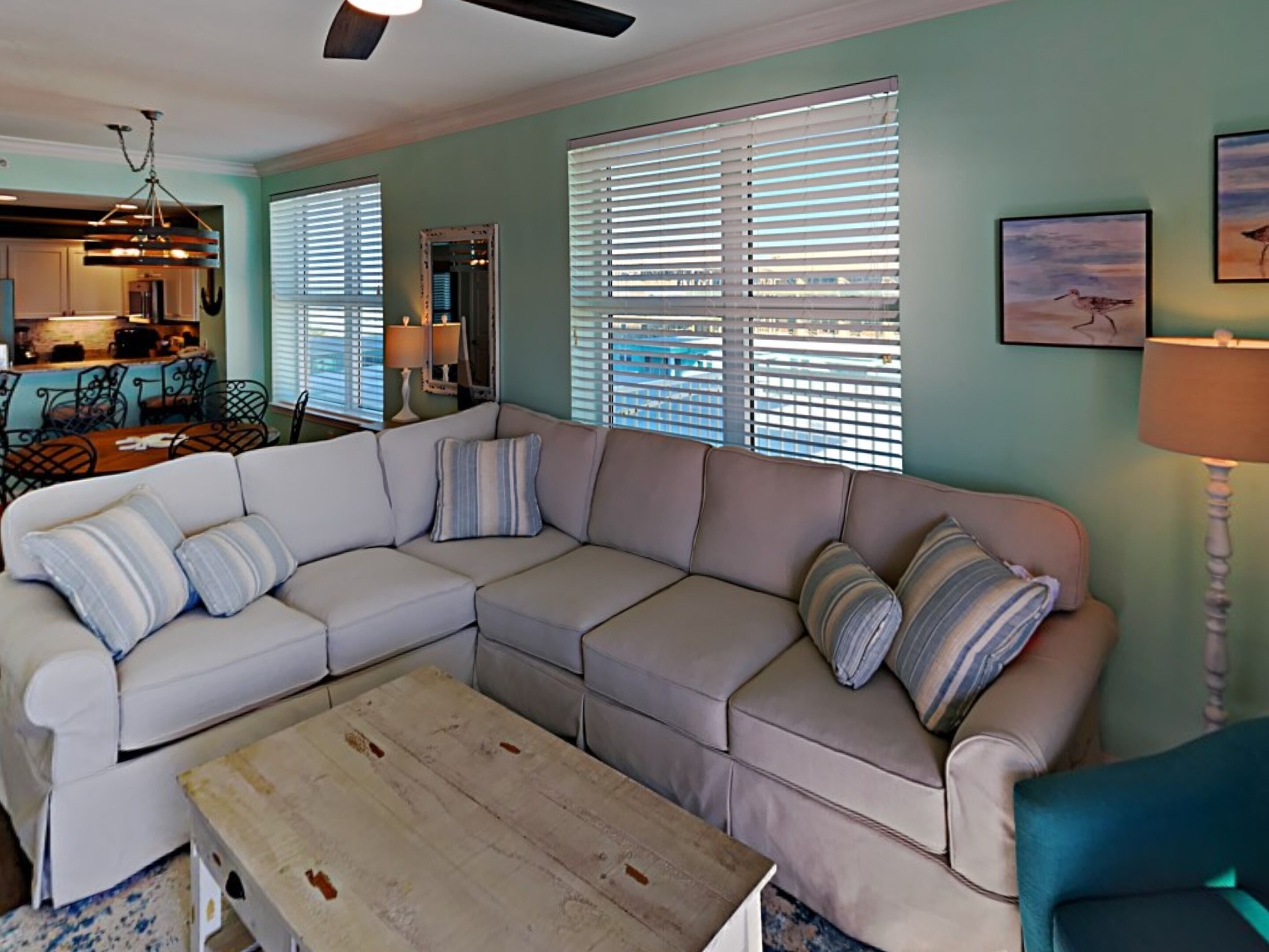 A living room with a beige sectional sofa, a wooden coffee table, and a teal armchair. There are two windows with blinds, a ceiling fan, and a kitchen area in the background.
