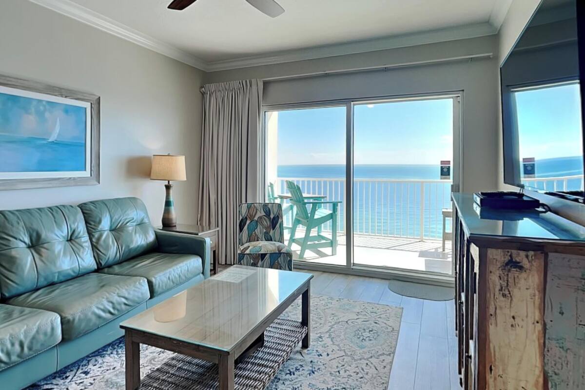 A spacious living room with a large window offering a view of the ocean, a ceiling fan, a leather sofa, a coffee table, and a television.