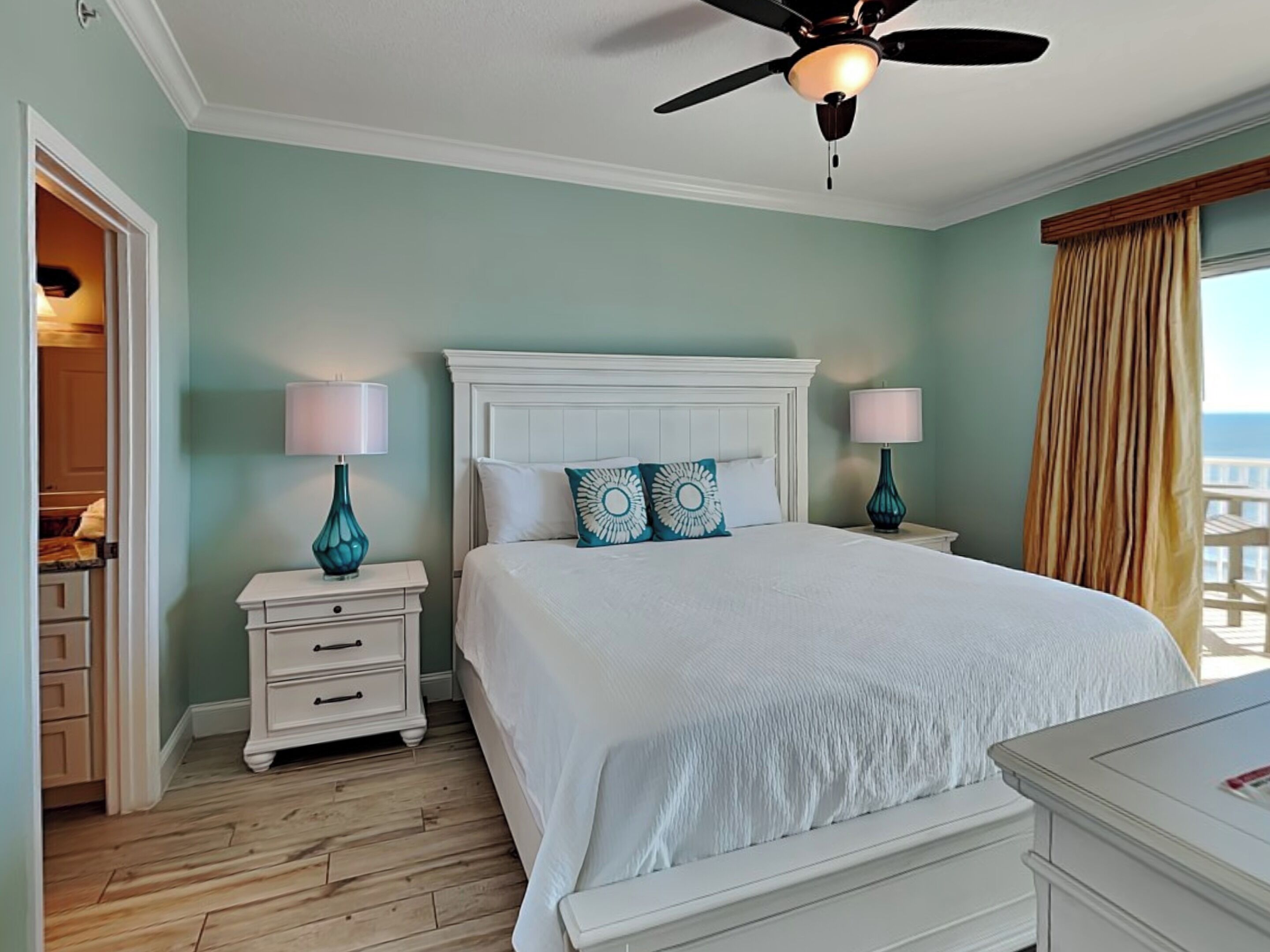 A bedroom with a white bed, two bedside tables with lamps, a ceiling fan, and a view of the ocean through a window with curtains.