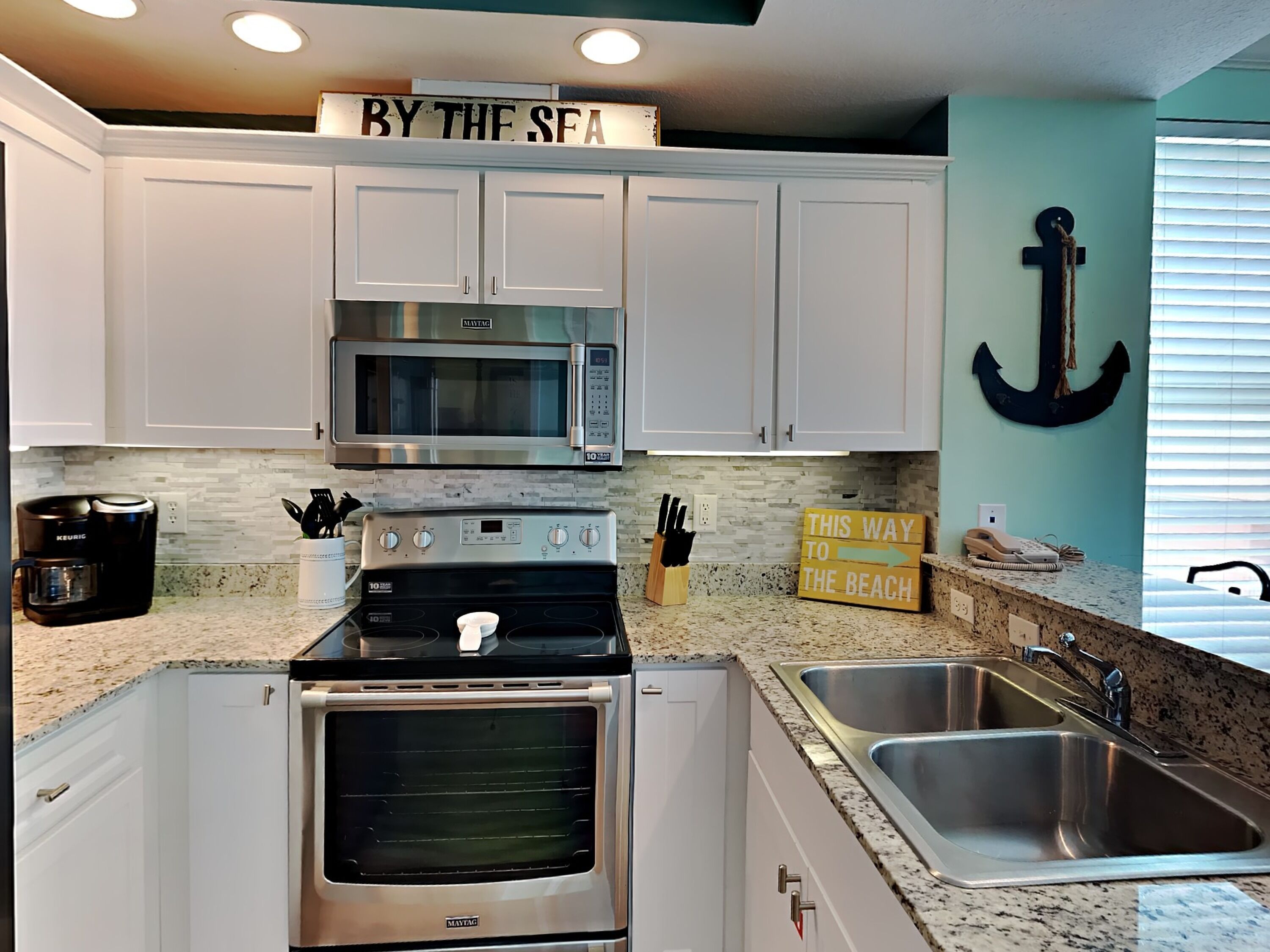 A kitchen with white cabinets, a Maytag oven, a stainless steel sink, and a sign that reads "BY THE SEA".