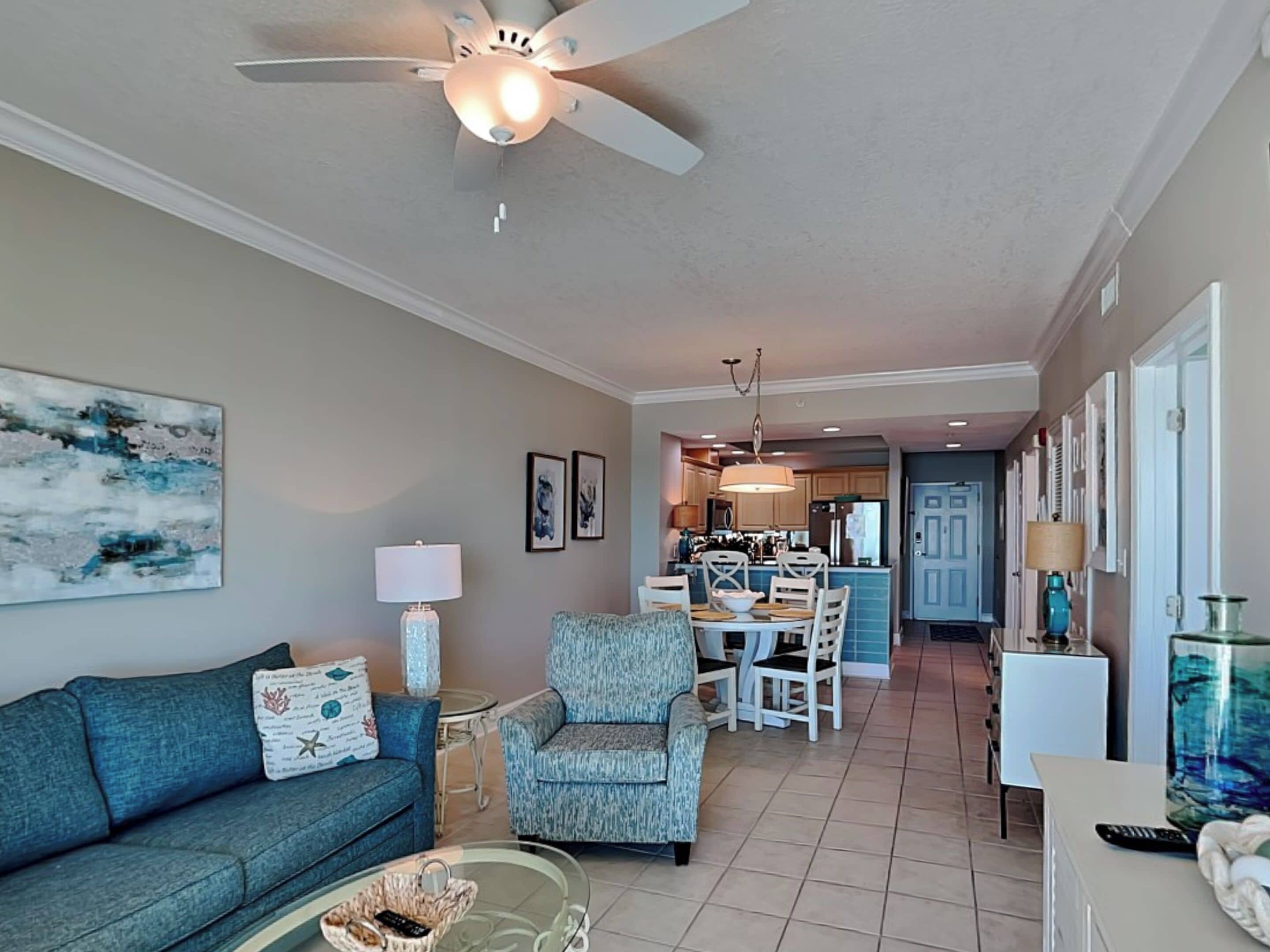 A living room with a sofa, armchair, and coffee table. There is a dining area with a table and chairs, and a kitchen in the background.