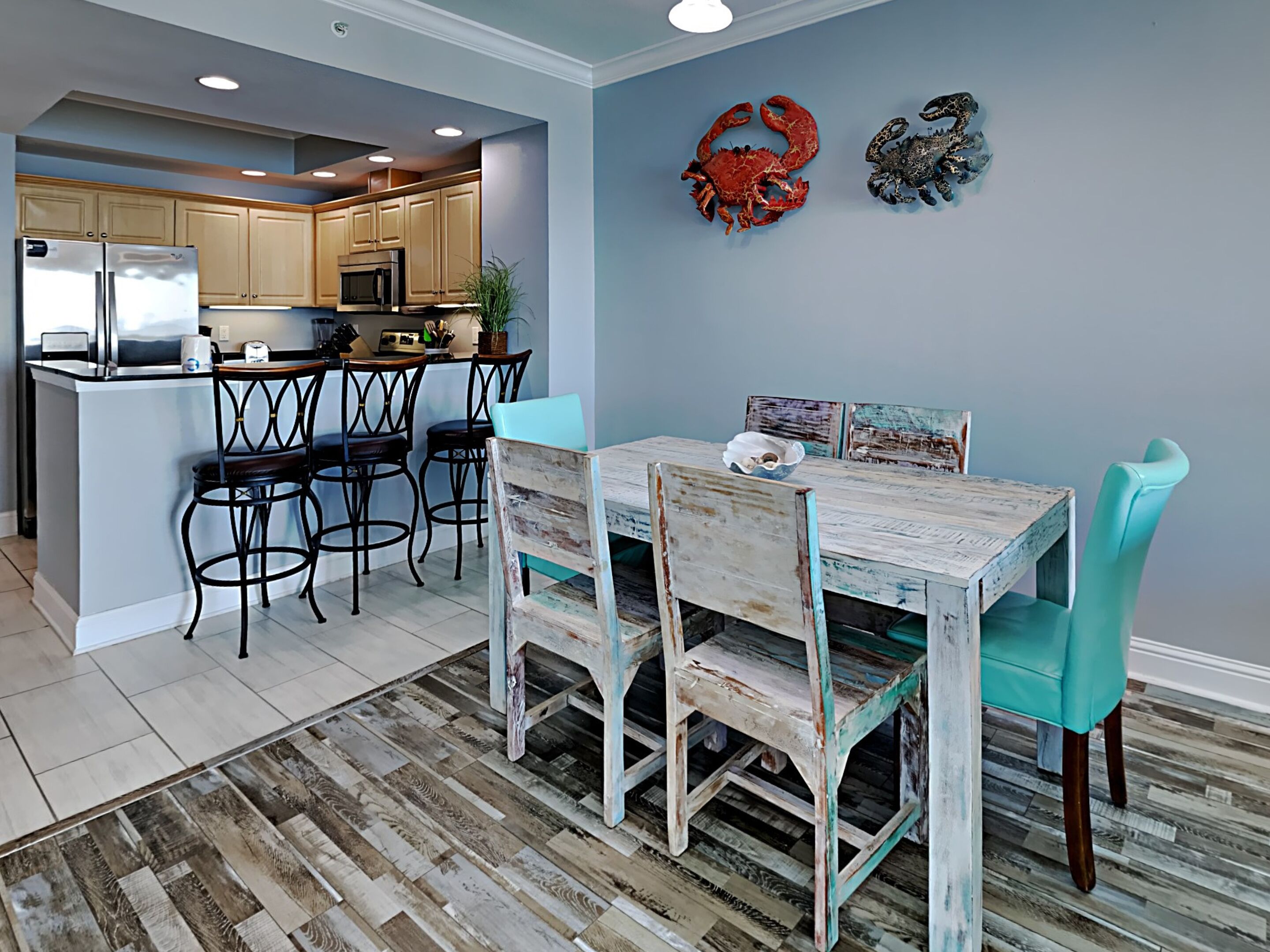 A dining area with a rustic wooden table and chairs, a kitchen in the background, and decorative crab wall art.