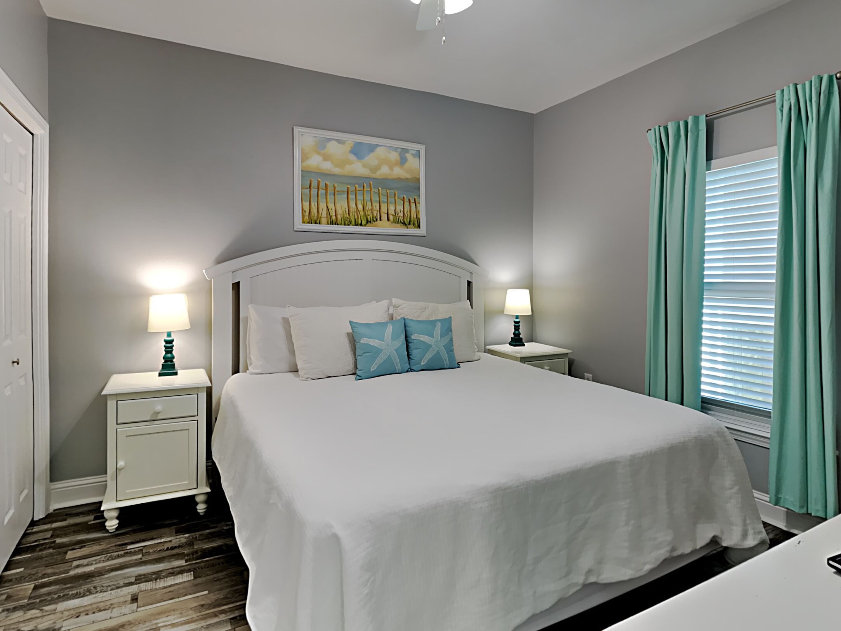 A bedroom with a large bed, two bedside tables, a ceiling light, a window with curtains, and a painting on the wall.