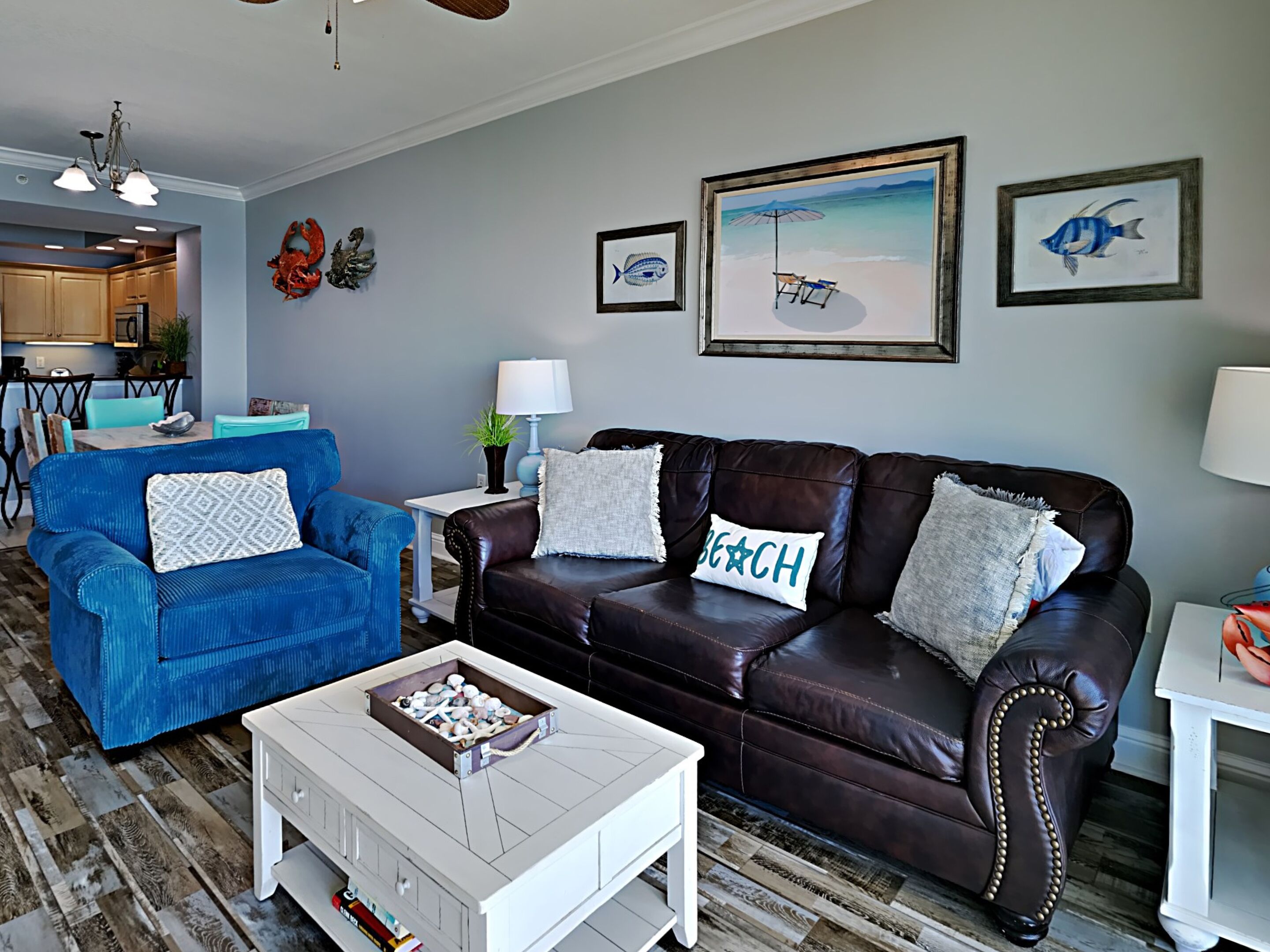 A living room with a blue armchair, a brown leather sofa, and a white coffee table. There are framed pictures on the wall and a chandelier above the seating area.