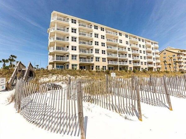 A multi-story hotel with balconies, located near a sandy beach with a wooden fence.
