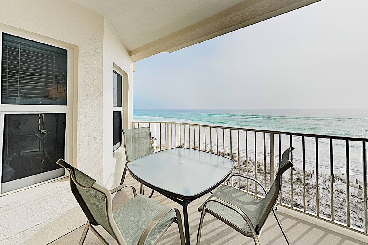 A balcony with a glass table and four chairs, overlooking a beach with the ocean and a clear sky.