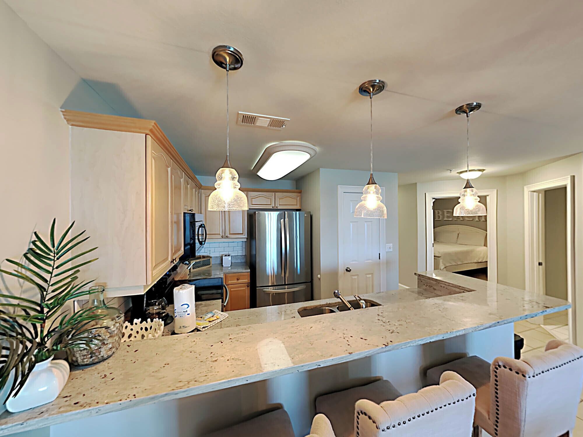 A modern kitchen with a large island, stainless steel appliances, pendant lighting, and a view into a bedroom.