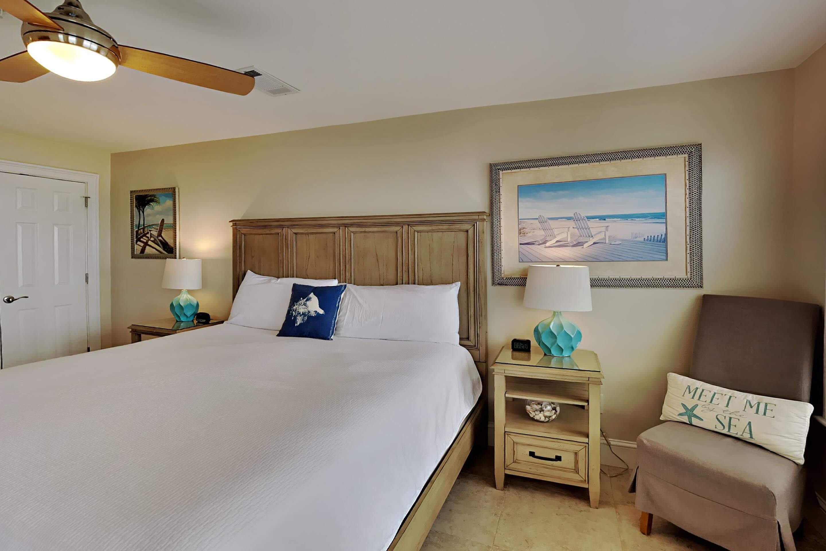 A neatly arranged hotel room with a large bed, a ceiling fan, a framed painting, and a chair with a pillow.