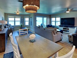 A spacious living area with a wooden dining table, sofas, a TV, and large windows offering a view of the outdoors.