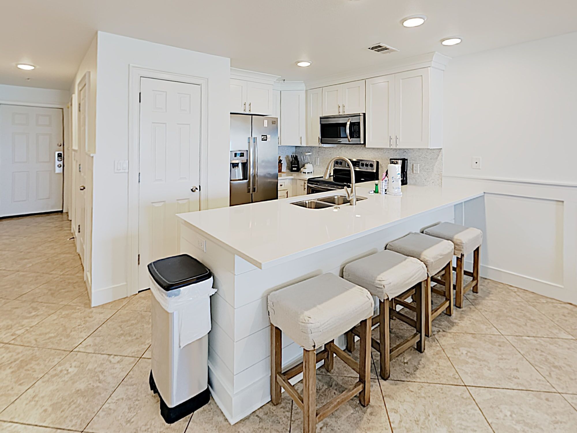 A modern kitchen with white cabinetry, a stainless steel refrigerator, a microwave, and a sink. There are four bar stools with cushioned seats.