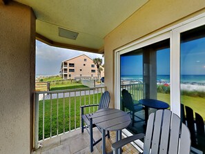 A balcony with a table and two chairs, overlooking a beach and a building.