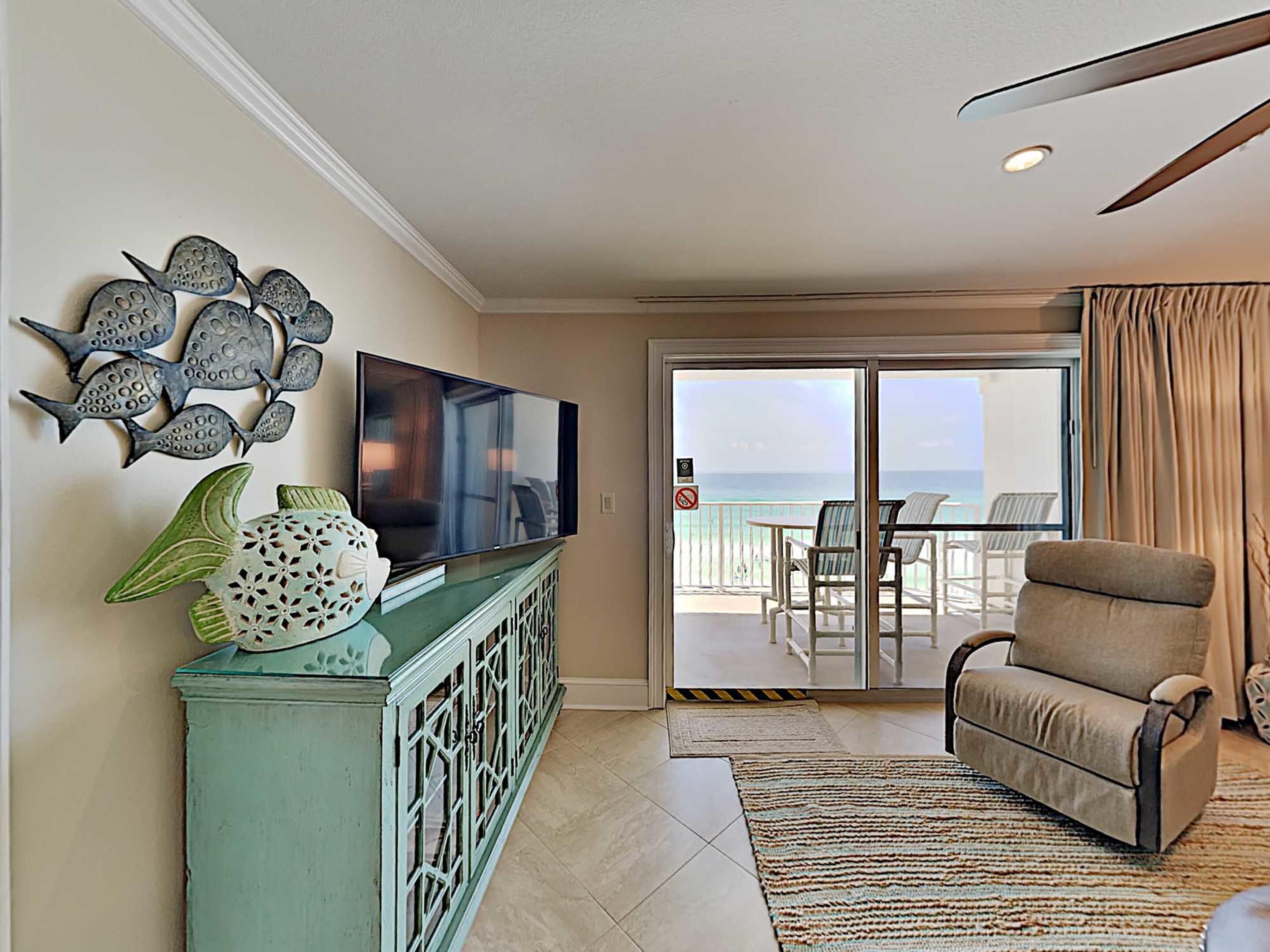 A living room with a TV, decorative fish wall art, a green cabinet, a beige armchair, and a view of the ocean through a sliding glass door.
