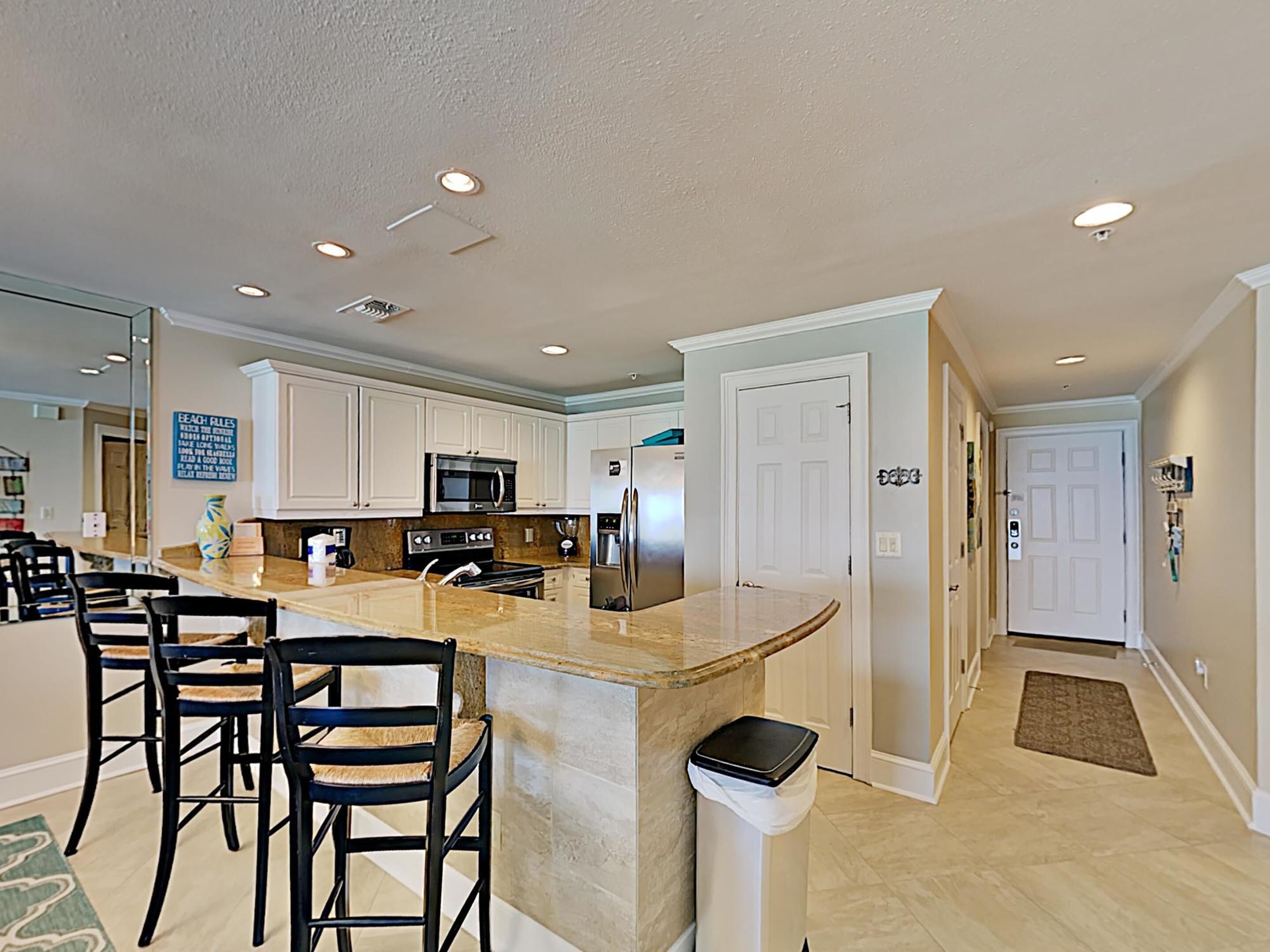 A modern kitchen with white cabinets, a granite countertop, a stainless steel refrigerator, and a microwave. There are three bar stools and a trash can.