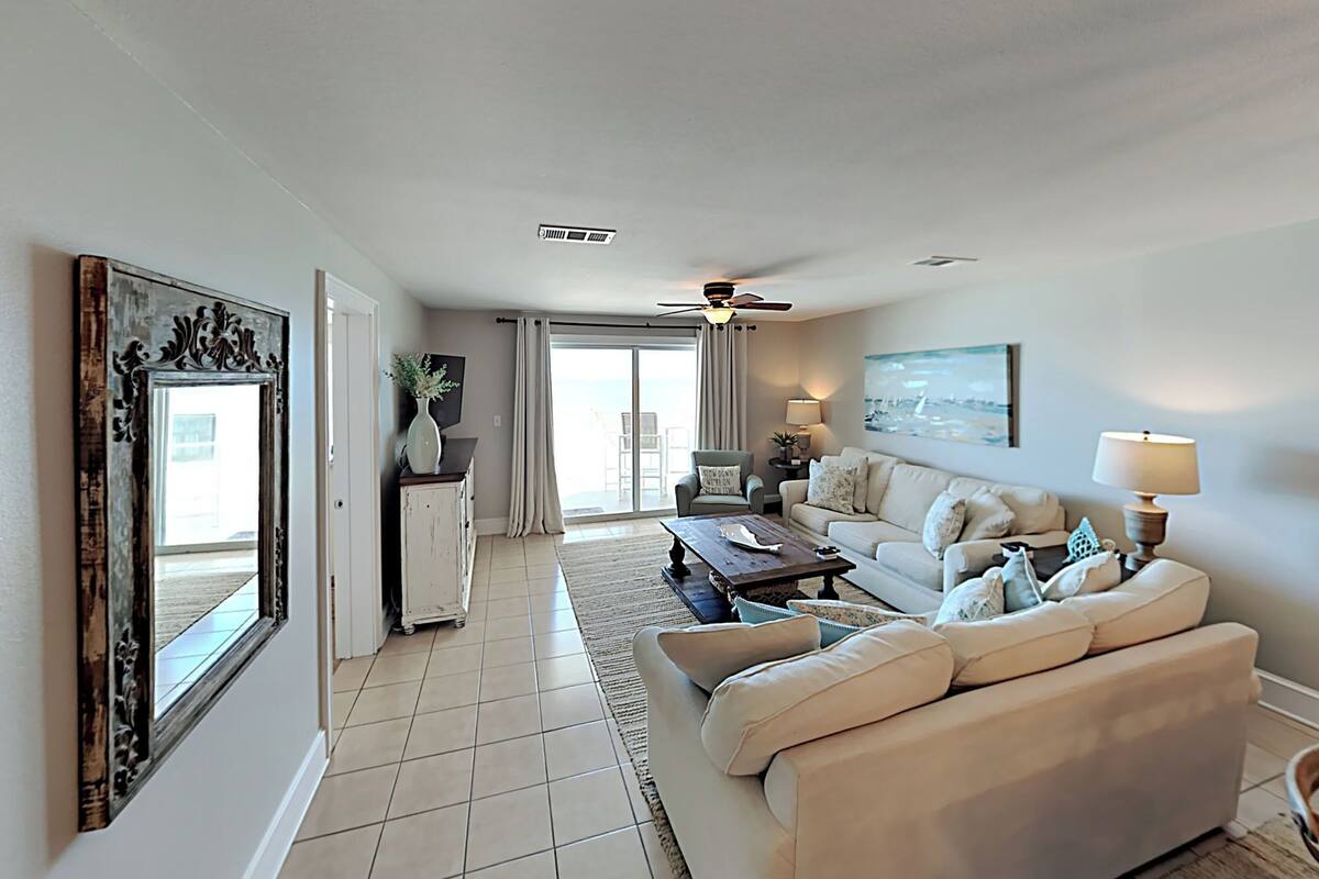 A spacious living room with a large sofa, a coffee table, and a television. There is a sliding glass door leading to a balcony.