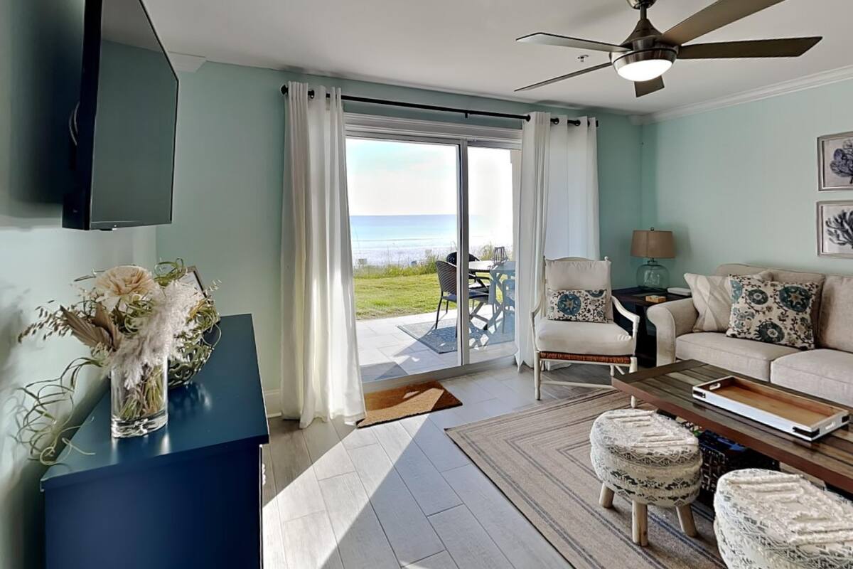 A living room with a view of the sea, a ceiling fan, a sofa, a coffee table, and a TV mounted on the wall.