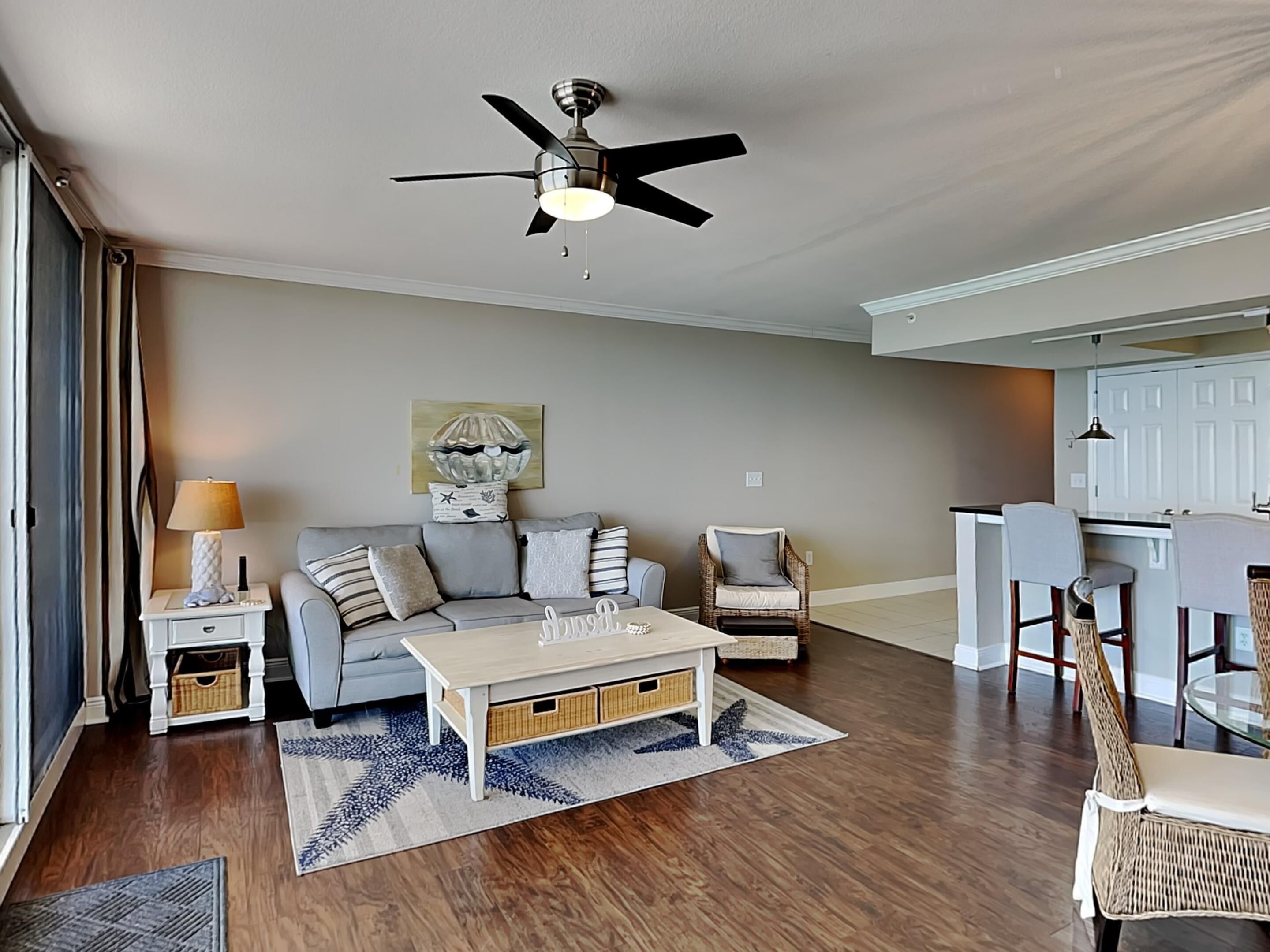 A modern living room with a sofa, coffee table, and chairs, a dining area with a kitchen in the background, and a ceiling fan.