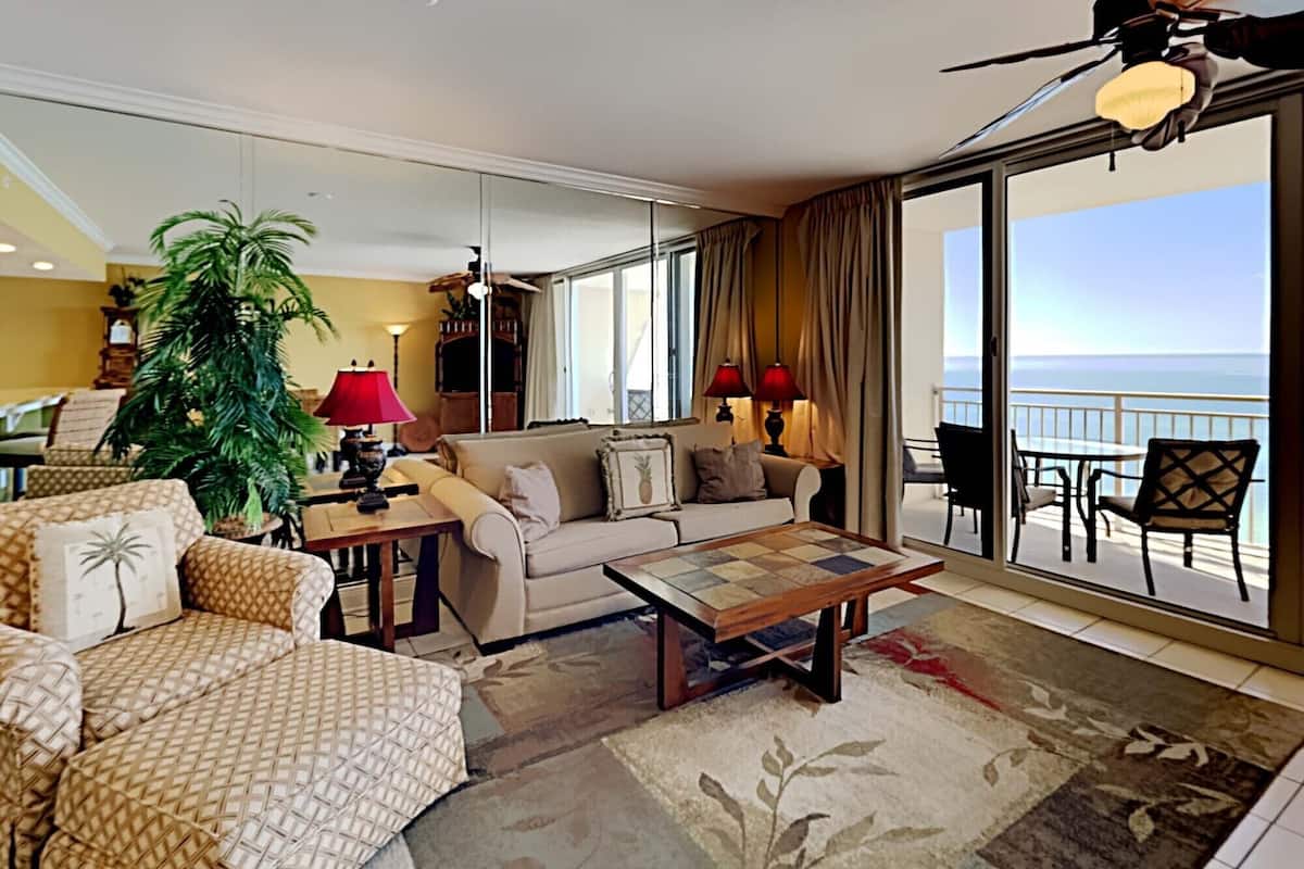 Uma sala de estar ampla com uma grande janela que oferece vista para o mar, um sofá, uma mesa de centro e um vaso com planta.