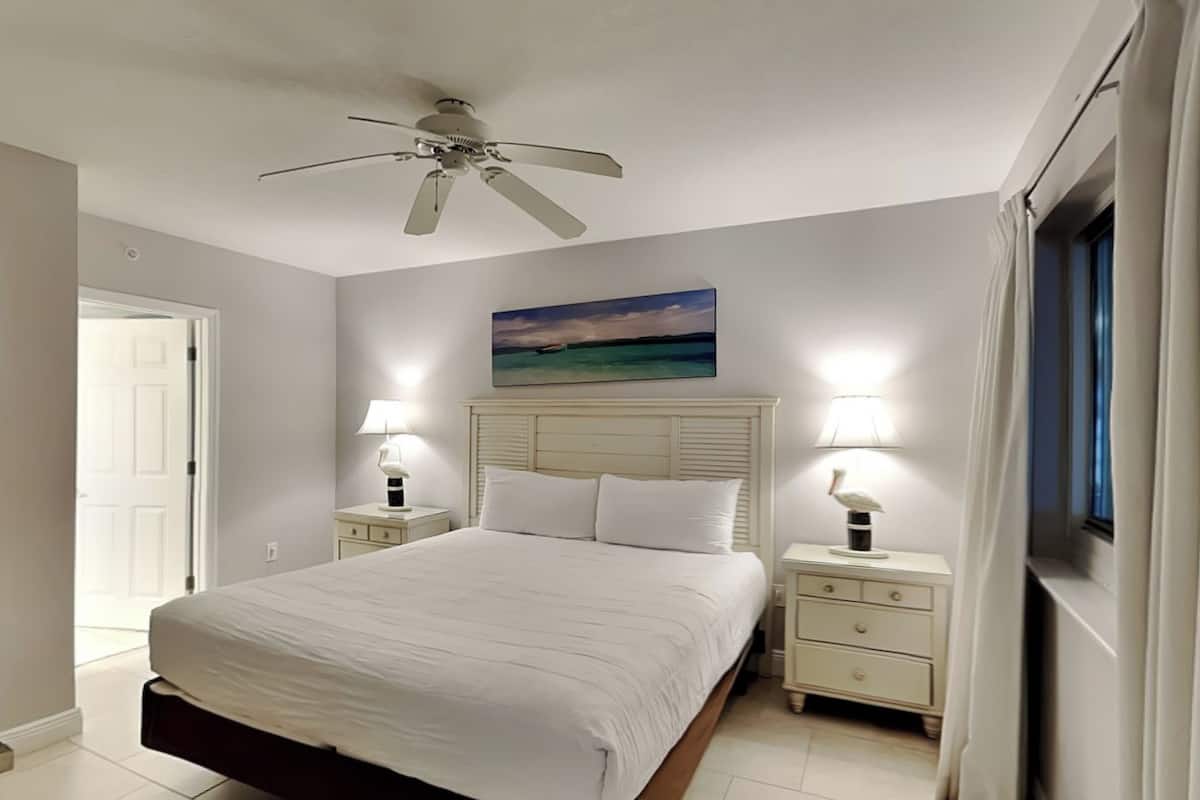 Um quarto com uma cama, duas mesas de cabeceira com abajures, um ventilador de teto e uma pintura na parede.
