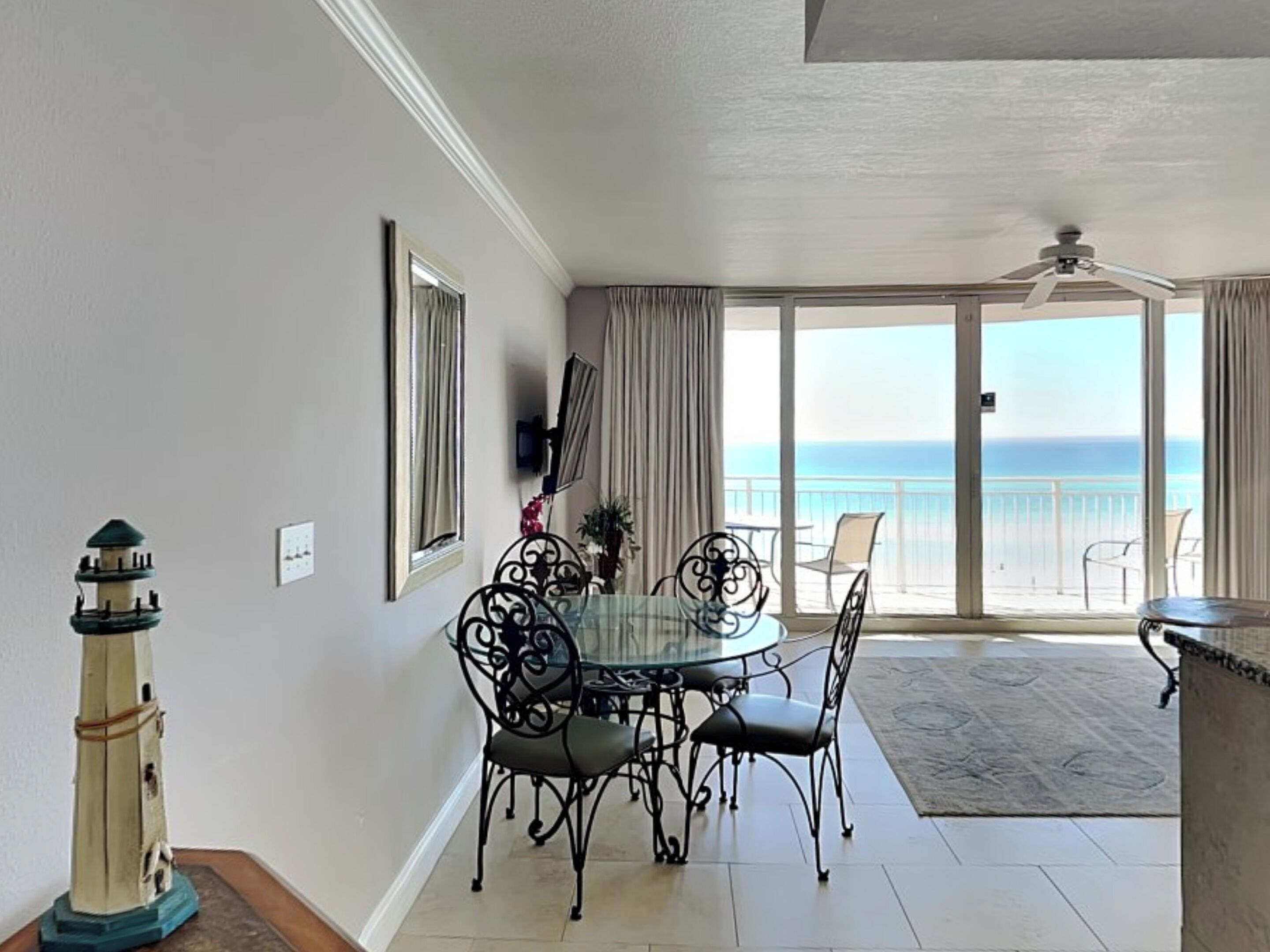 A dining area with a round table and chairs, a television on the wall, and a view of the ocean through large sliding doors.