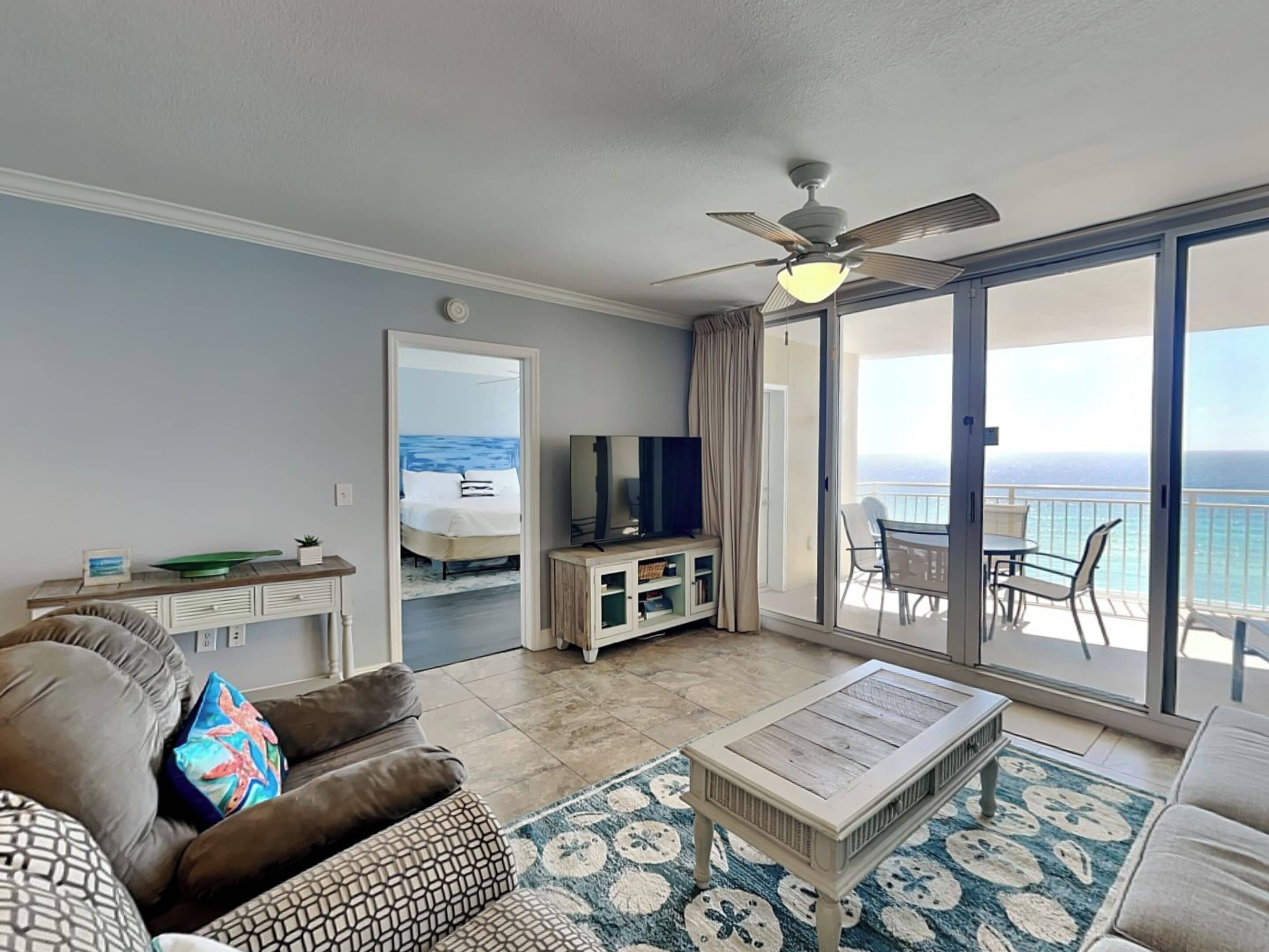 A living room with a sofa, coffee table, TV, and a balcony with a view of the ocean.