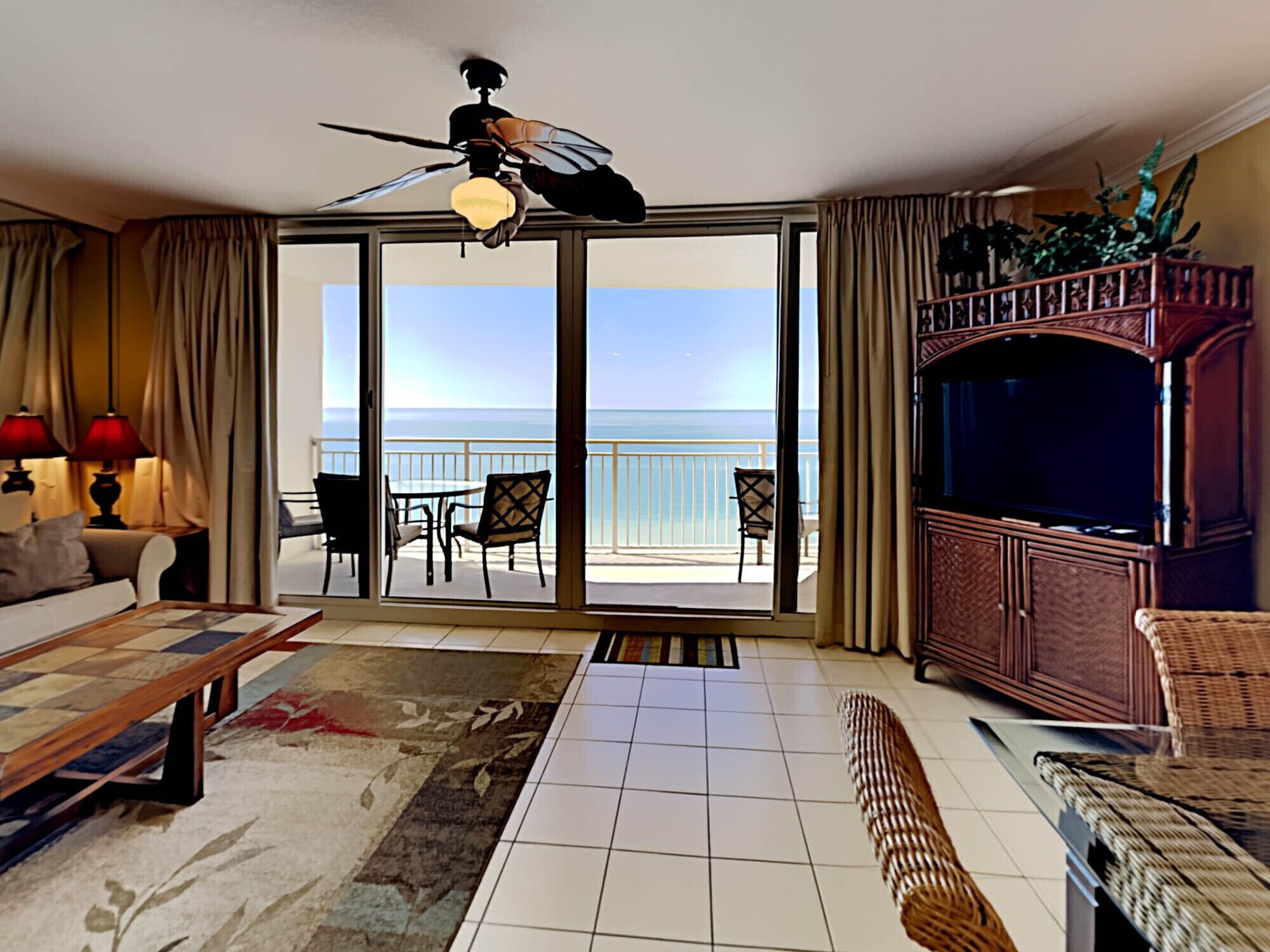 A spacious living room with a large window overlooking the sea, a ceiling fan, a television, and a dining area with a table and chairs.