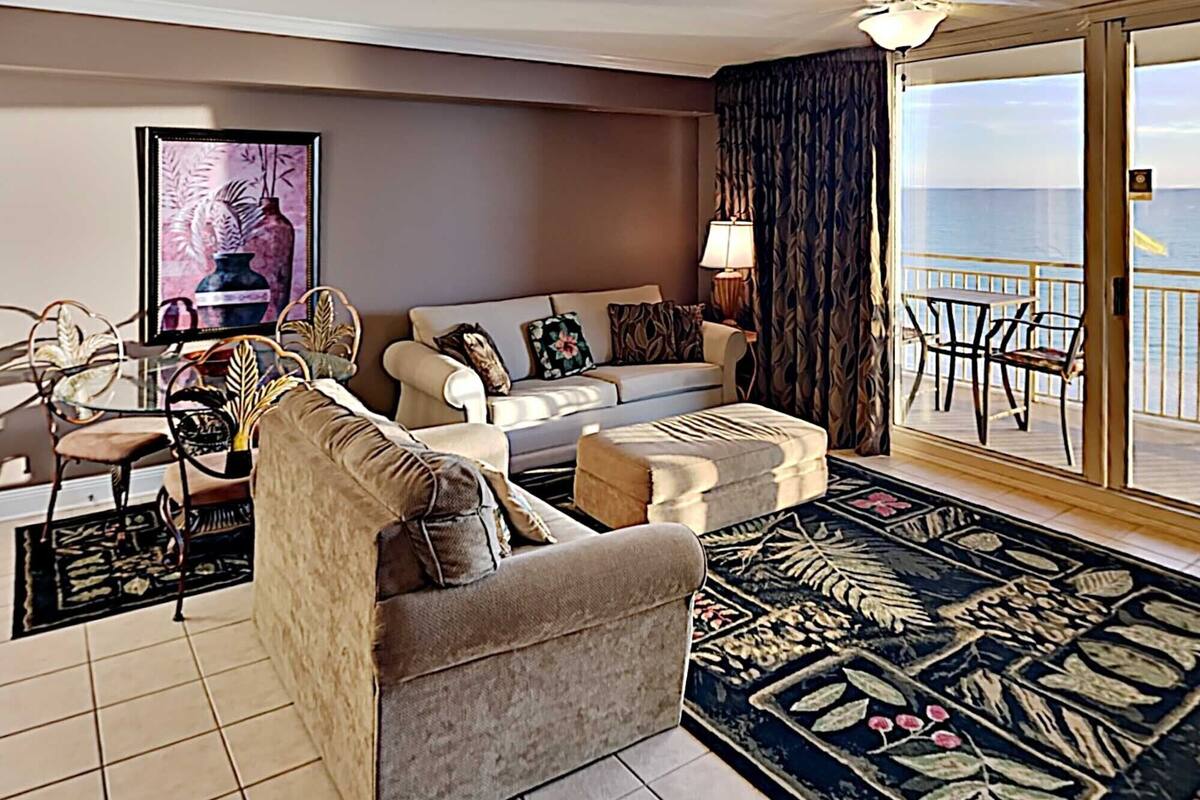 Uma sala de estar com sofá, poltrona e um banquinho, uma área de jantar com cadeiras, uma mesa e uma grande janela com vista para o mar.