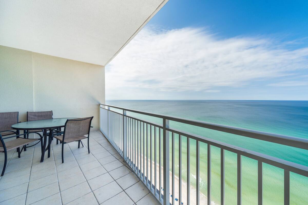 Varanda com mesa e cadeiras, vista para um mar azul-turquesa e céu claro.