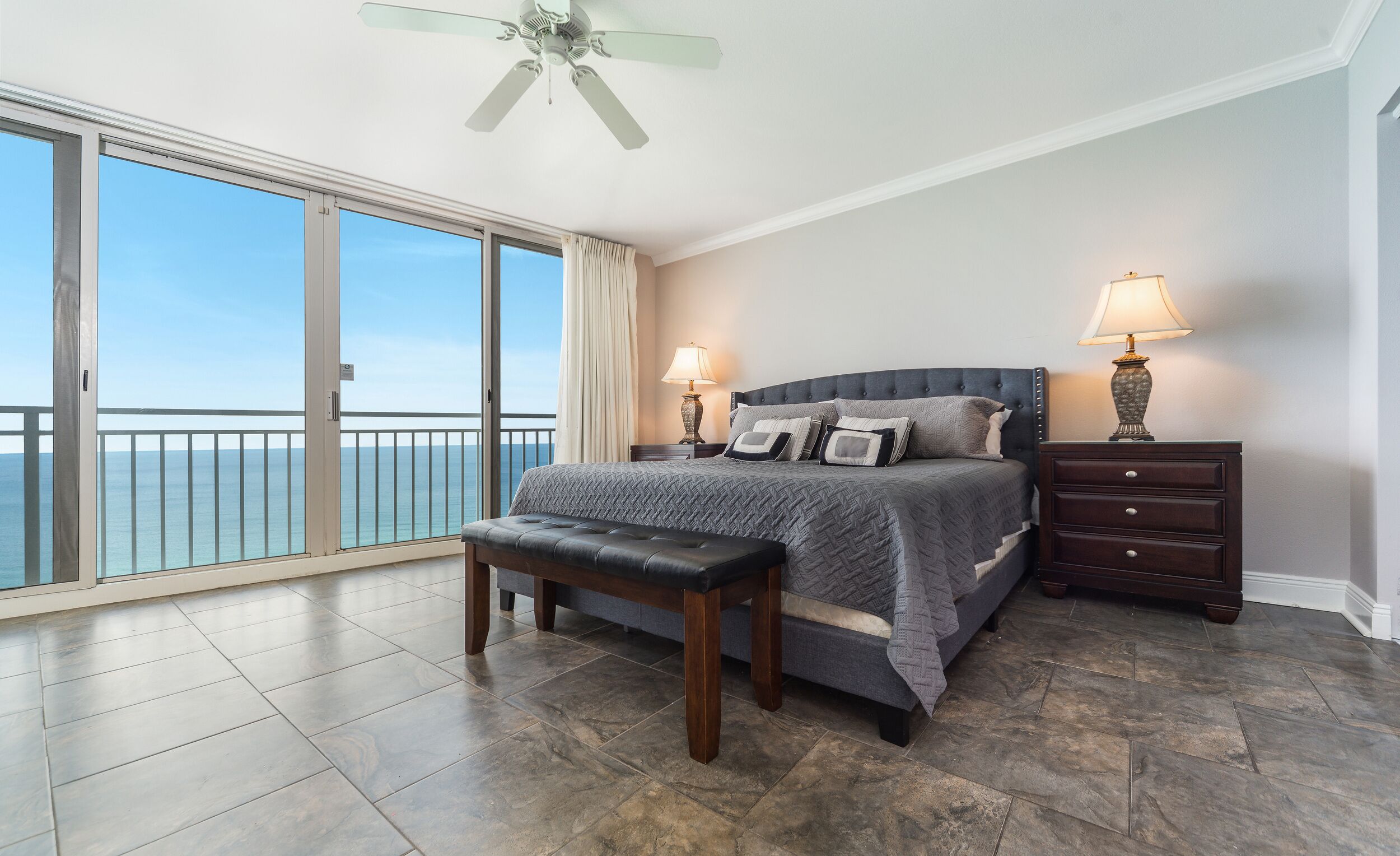 A bedroom with a large bed, two bedside tables, a bench, a ceiling fan, and a view of the ocean.