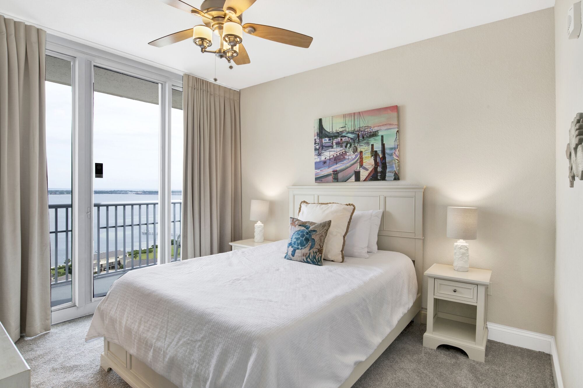 Una habitación de hotel con una cama grande, un ventilador de techo, un cuadro que muestra un puerto y vistas al agua a través de las puertas del balcón.