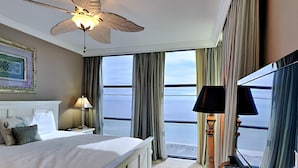 Une chambre d’hôtel avec un lit, un ventilateur de plafond, une grande fenêtre avec des rideaux, un piano et un tableau encadré au mur.