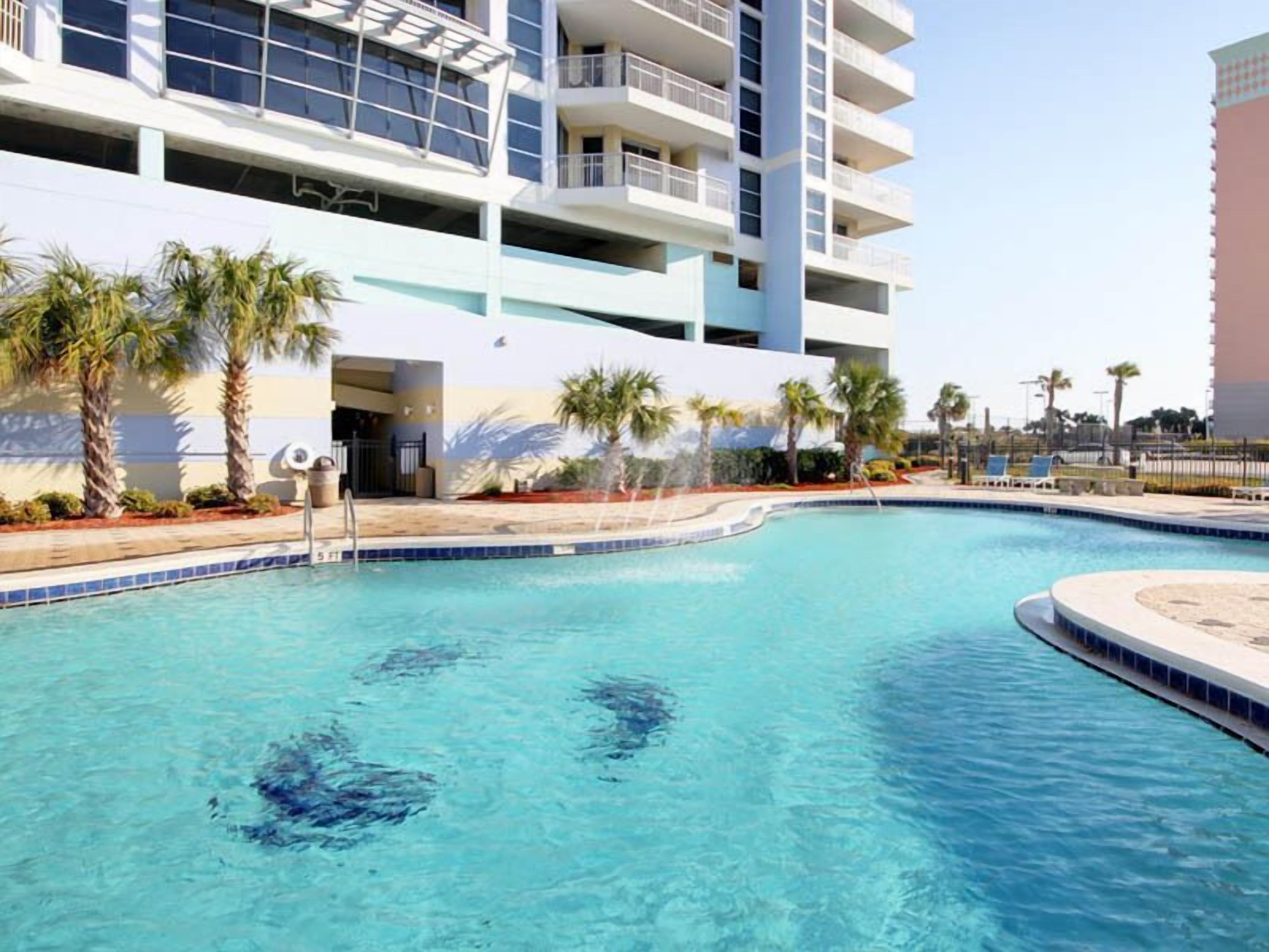 Una piscina con agua cristalina y azul, rodeada de palmeras y un edificio de varios pisos con balcones.