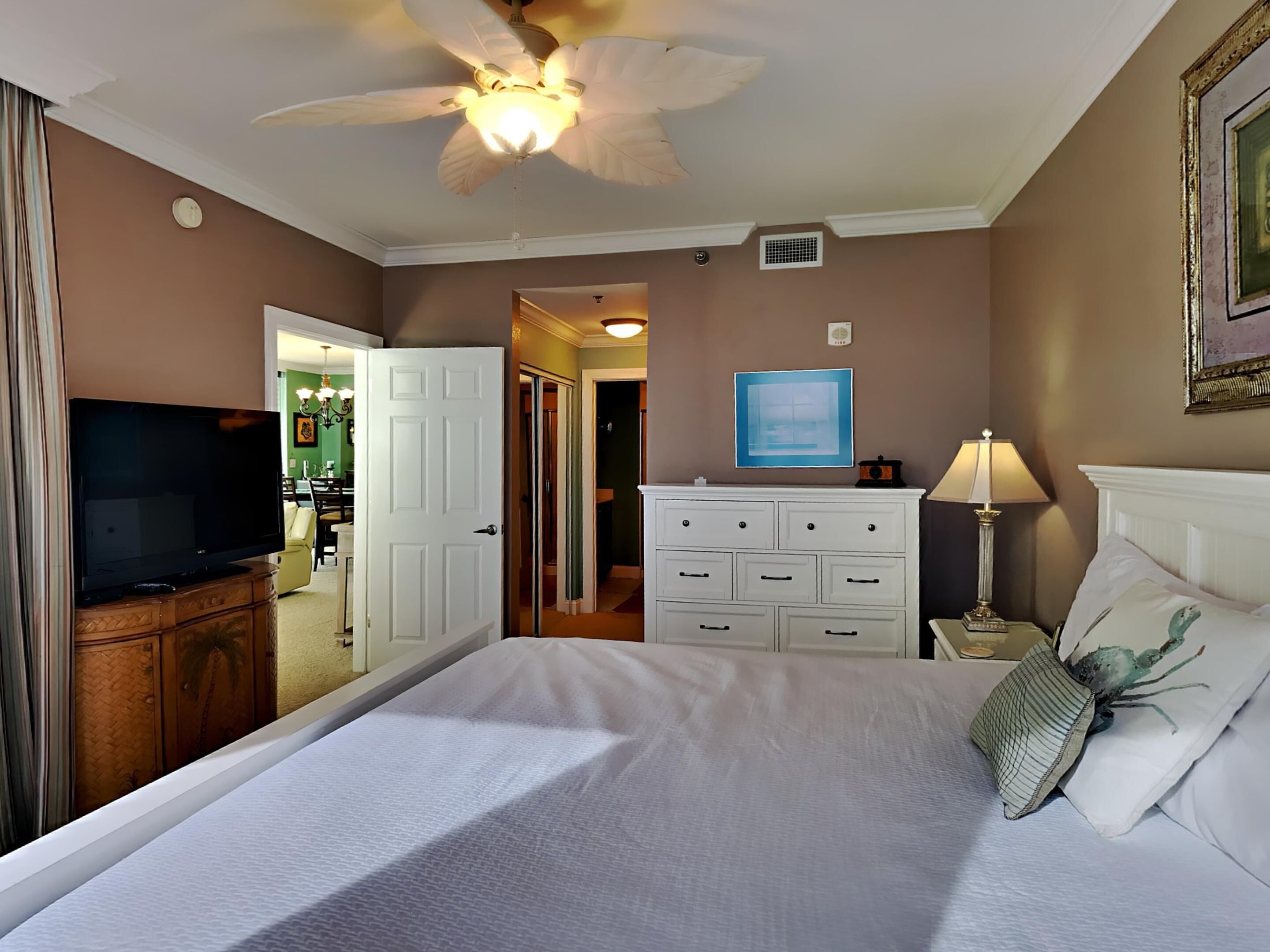 A hotel room with a bed, a television, a dresser, a ceiling fan, and a door leading to another room.