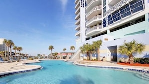 Un espace piscine avec une eau d’un bleu limpide, des palmiers et un immeuble à plusieurs étages doté de balcons.