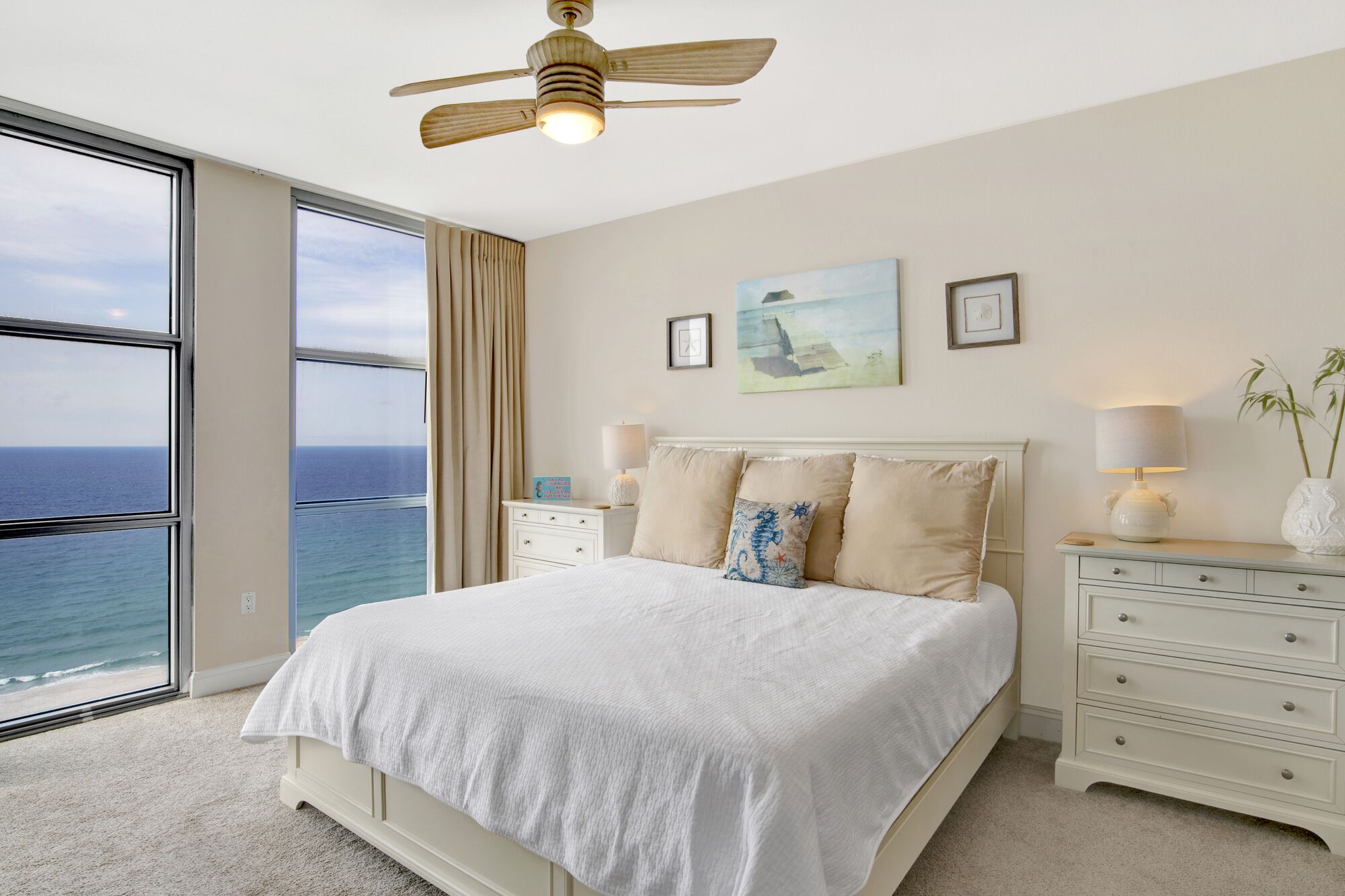A bedroom with a large bed, two bedside tables, a ceiling fan, and a view of the ocean.