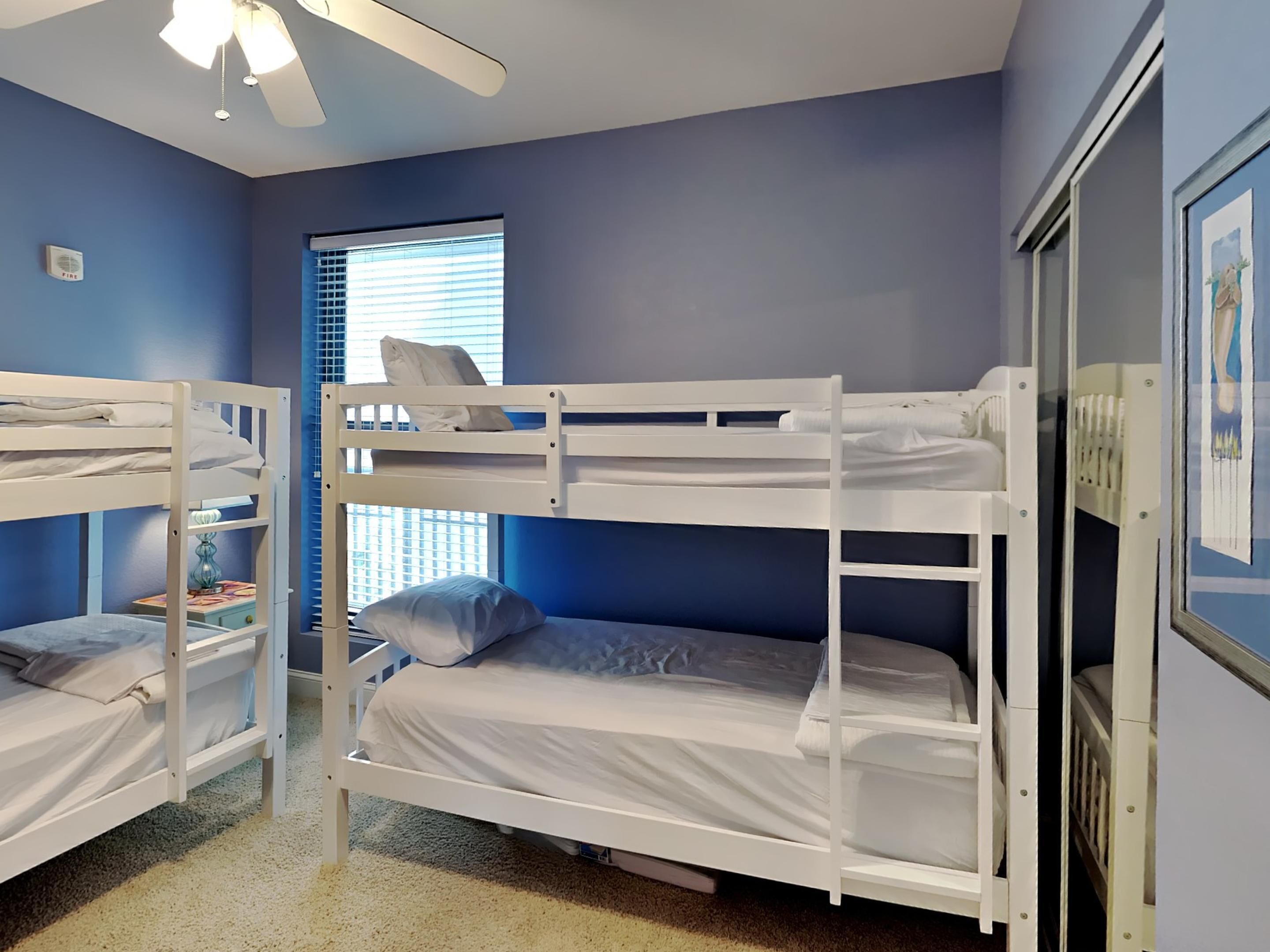 A bunk bed room with a ceiling fan, a window with blinds, and a framed picture on the wall.