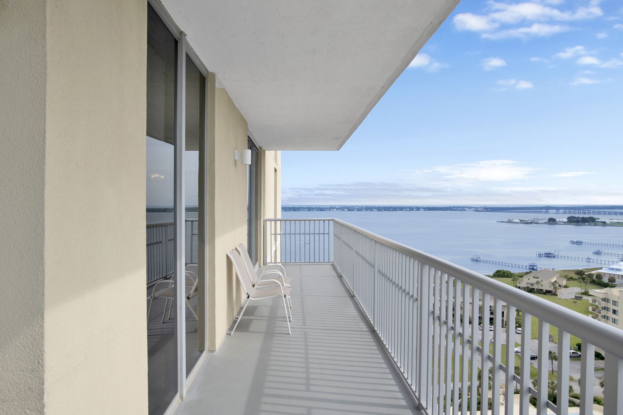 A balcony with white chairs, a view of the water and a bridge, and a clear sky.