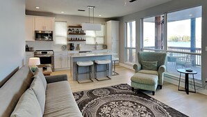 A modern kitchen with white cabinets, a central island, and bar stools. There is a grey armchair and a patterned rug in the living area. - Destiny Beach Villas by Southern Vacation Rentals (Destin)
