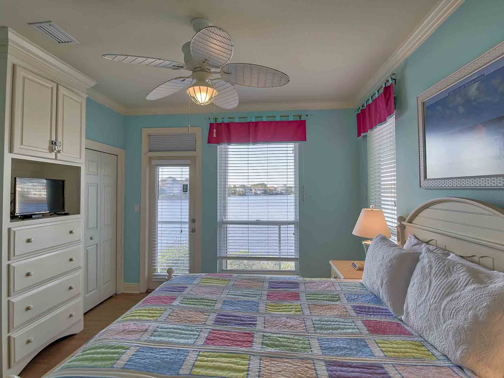 A bedroom with a bed, a ceiling fan, a television on a dresser, and a window with blinds.