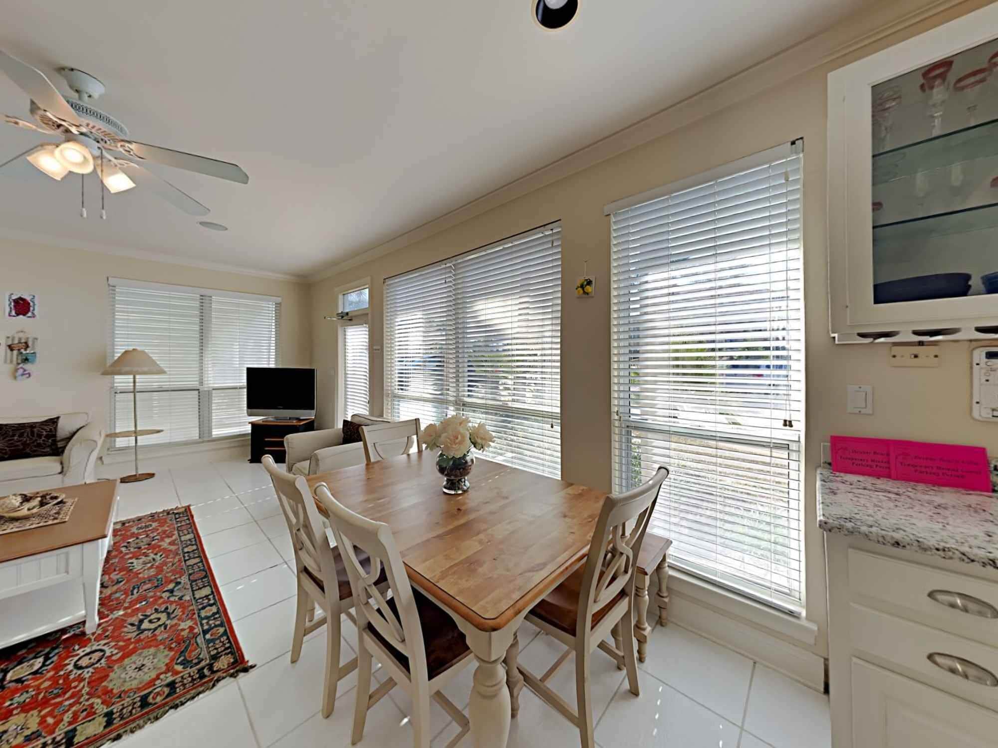 A spacious living room with a wooden dining table, white chairs, a ceiling fan, and large windows with blinds.