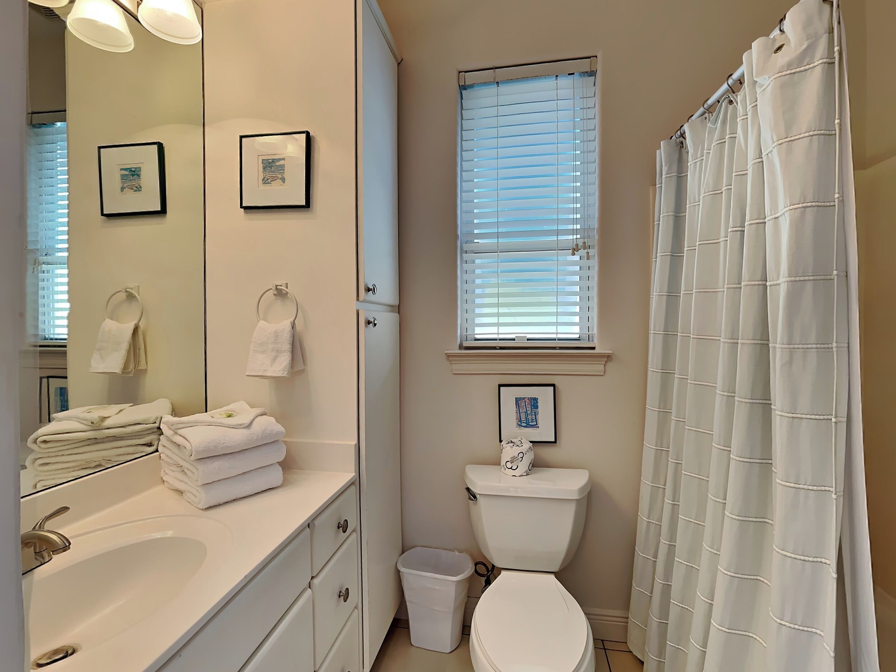 A bathroom with a sink, toilet, and shower curtain. There are towels stacked on the counter and a trash bin next to the toilet.
