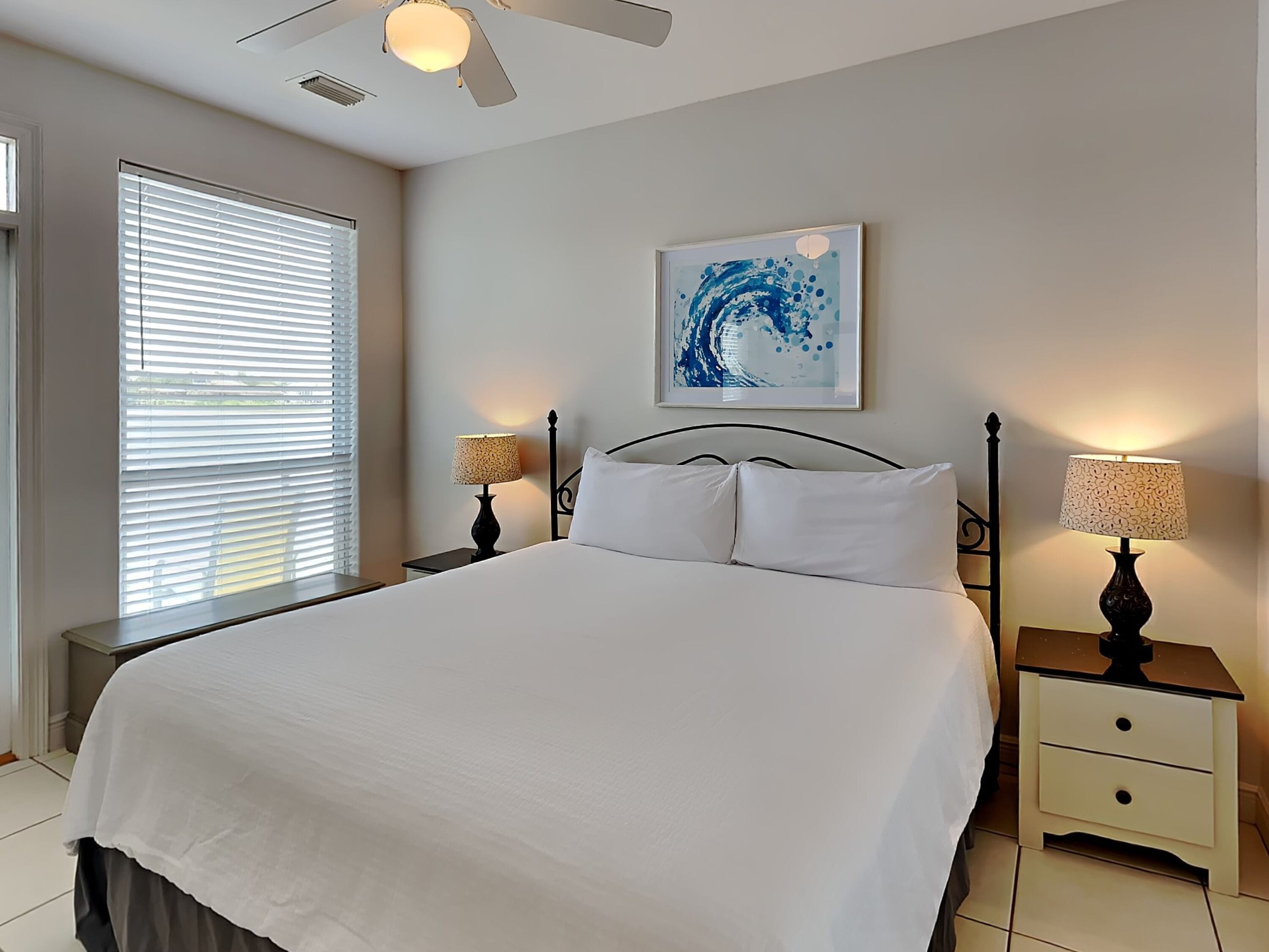 A bedroom with a bed, two bedside tables, a ceiling fan, a window with blinds, and a wall-mounted abstract painting.