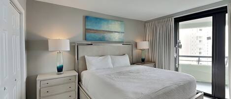 Quarto de hotel com uma cama grande, duas mesas de cabeceira com abajures, um quadro na parede e vista através de uma janela ampla.