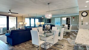 A modern living room with a glass dining table, white chairs, a blue sofa, and a chandelier. - Edgewater by Southern Vacation Rentals (Miramar Beach)