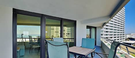 Un balcón con mesa y sillas, puertas de cristal amplias y vistas al océano y edificios.