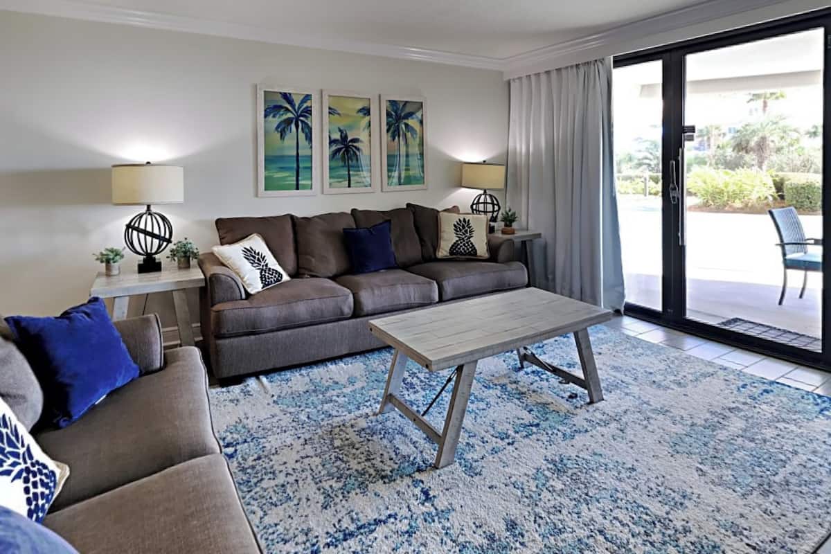 A living room with a sofa, coffee table, and two side tables. There are three framed artworks on the wall, a sliding glass door to the outside, and a view of a patio area.