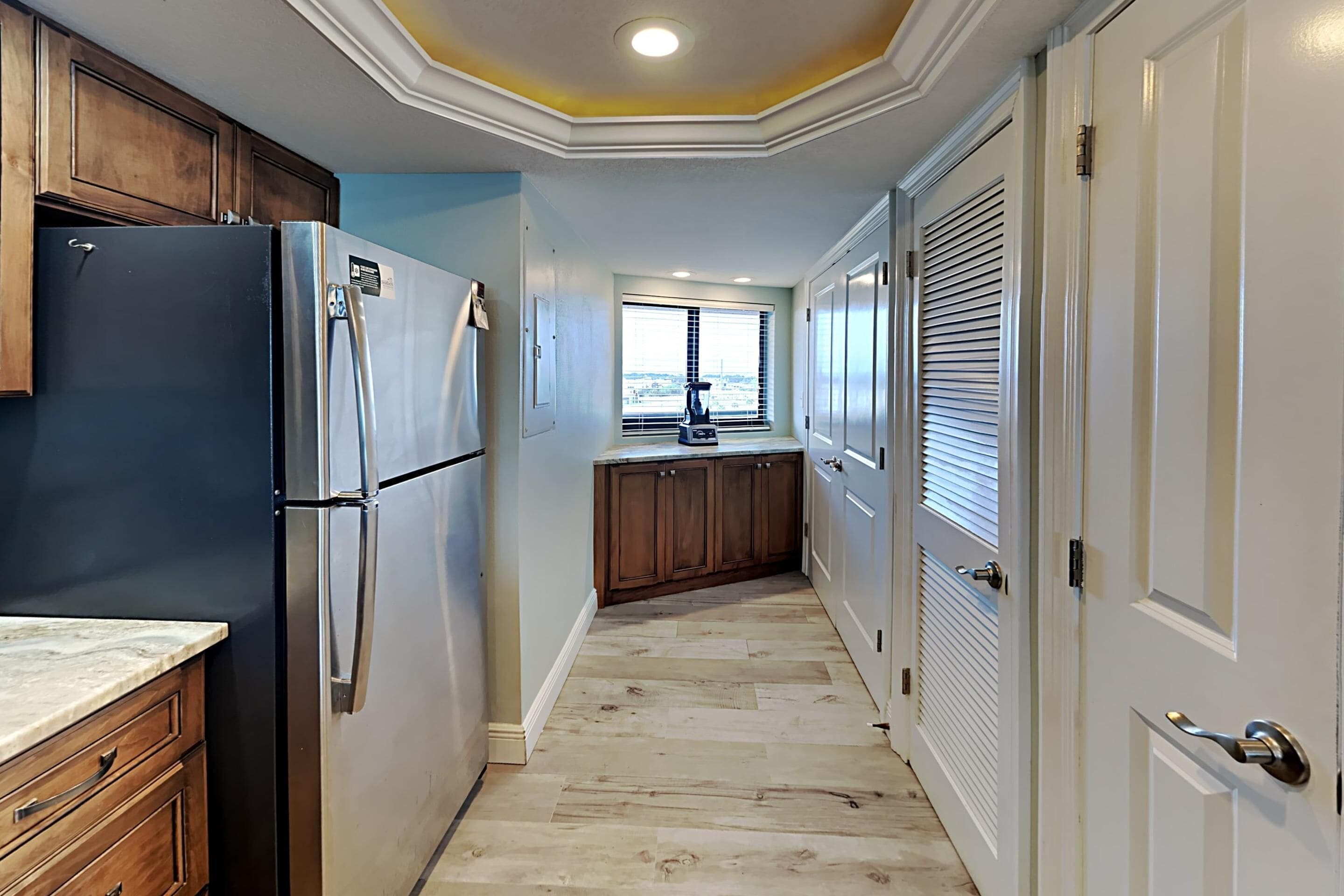 A narrow kitchen with a refrigerator, wooden cabinets, and a window with blinds.